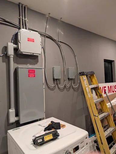 Electrical panel with red warning labels mounted on gray wall with ladder and equipment nearby